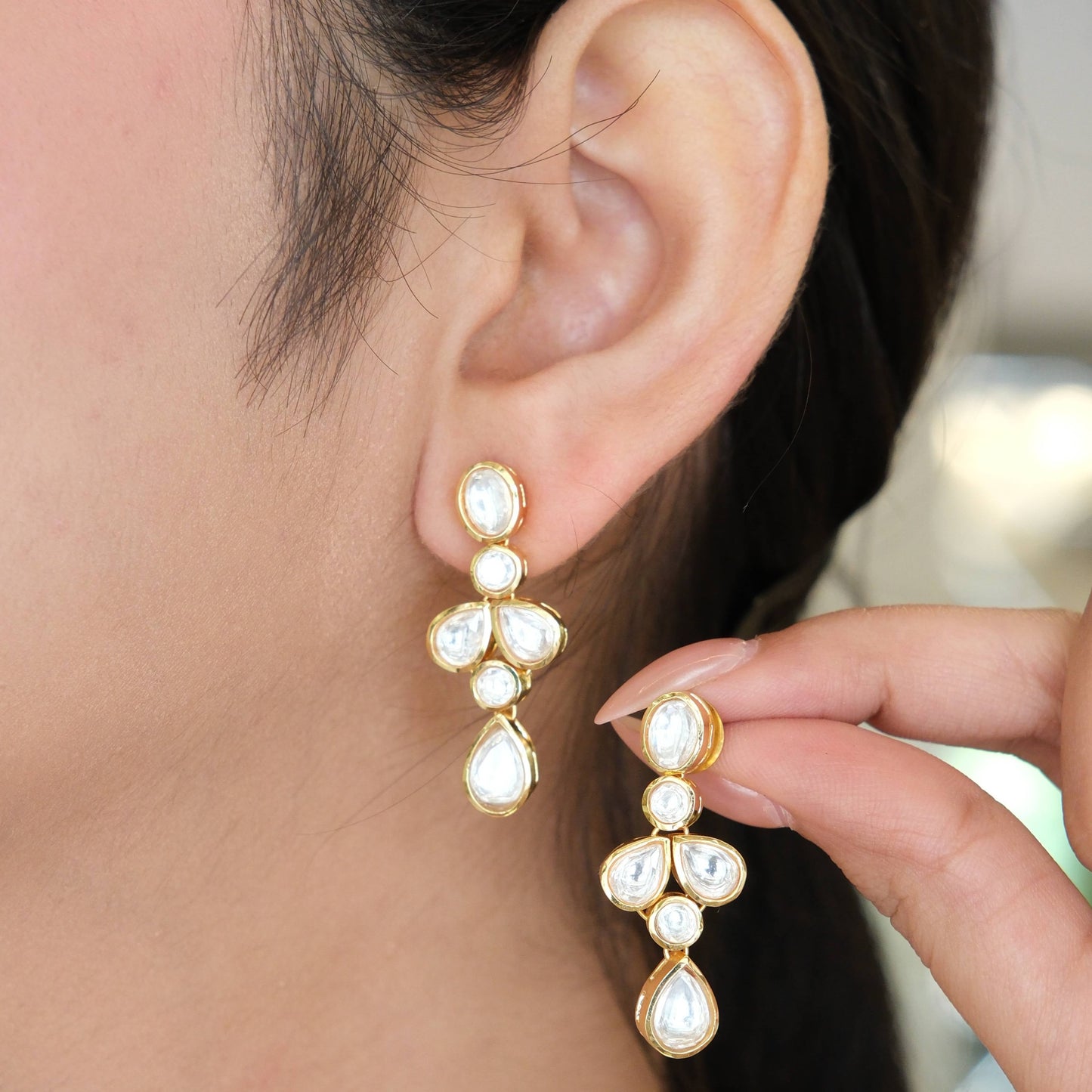 A close-up of a person wearing a gold and white drop earring while holding the matching earring in their hand.