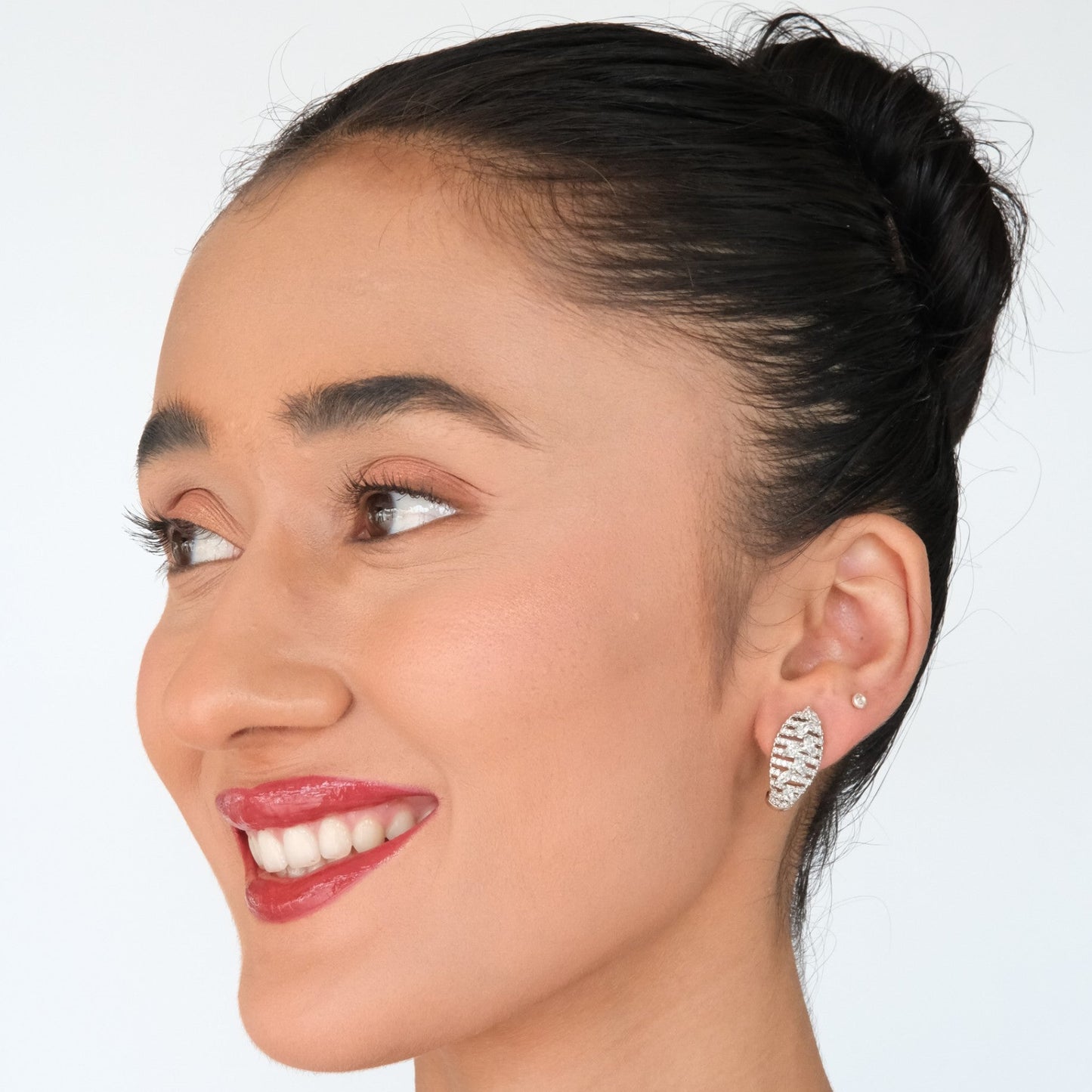 A close-up portrait of a smiling woman with dark hair in a bun, wearing a silver textured hoop earring on her left ear, against a plain light gray background.