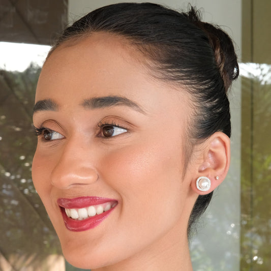 A close-up portrait of a smiling woman with dark hair pulled back, wearing a small round stud earring with a white pearl-like center and a gold-toned outer ring, set against an indoor background with soft window light.