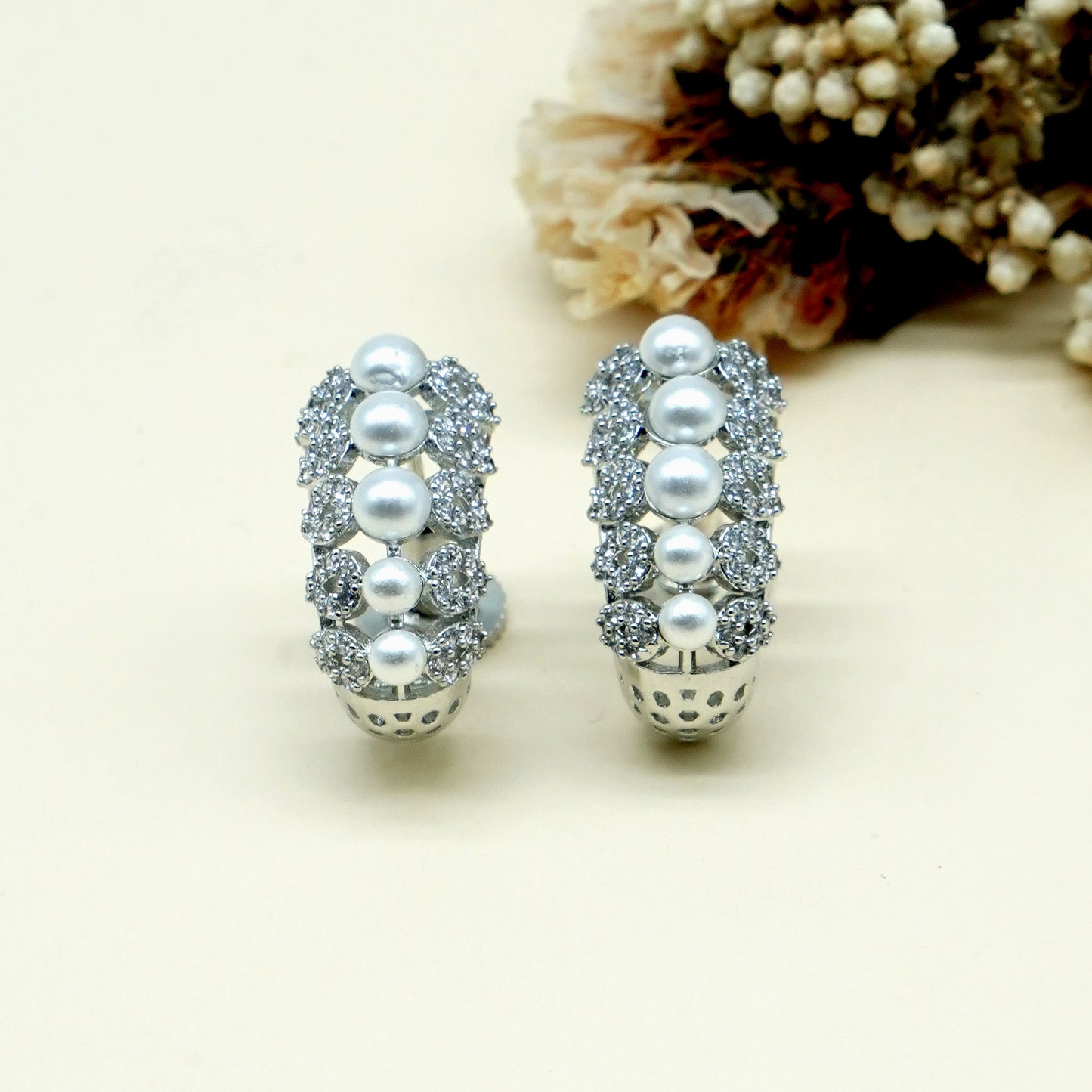 A pair of silver hoop earrings decorated with white pearls and clear rhinestones, displayed on a light cream background with dried flowers in the upper right corner.