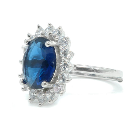 A silver ring featuring an oval-cut deep blue gemstone surrounded by a halo of small clear stones, set against a plain white background.