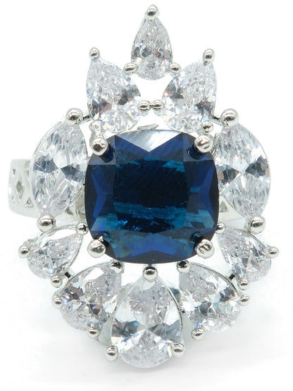 A silver-toned halo ring featuring a large central deep blue cushion-cut gemstone surrounded by a cluster of clear round and pear-cut stones, shown on a plain white background.