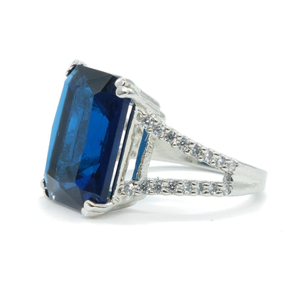 A silver ring featuring a large rectangular emerald-cut blue sapphire gemstone, accented with small pavé-set diamonds along the shoulders, shown on a plain white background.