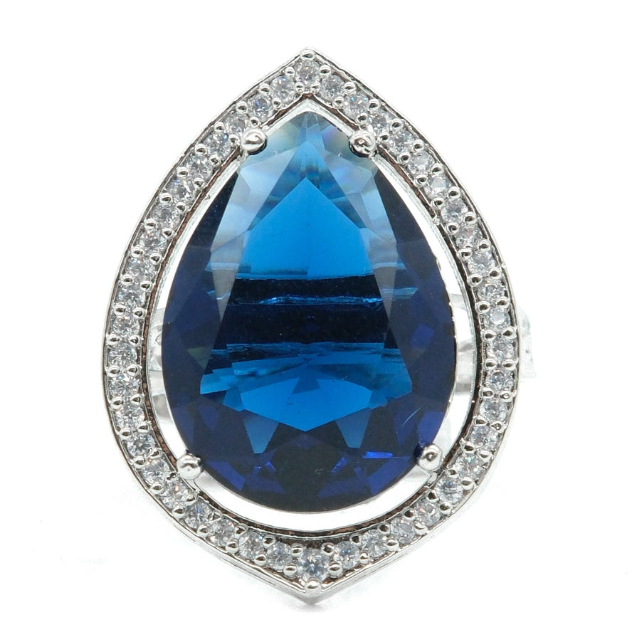 A teardrop-shaped silver ring featuring a large faceted blue gemstone in the center, surrounded by a halo of small clear stones, set against a plain white background.