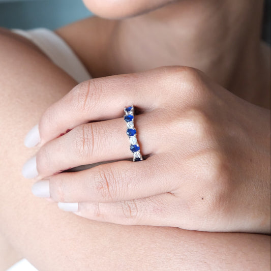 A close-up lifestyle shot of a woman's hand wearing a delicate silver band ring set with small, round blue gemstones.