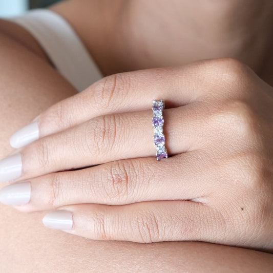 A close-up of a woman's hand wearing a slim silver band ring set with small lavender-purple gemstones, with her lips and shoulder also visible in the frame.