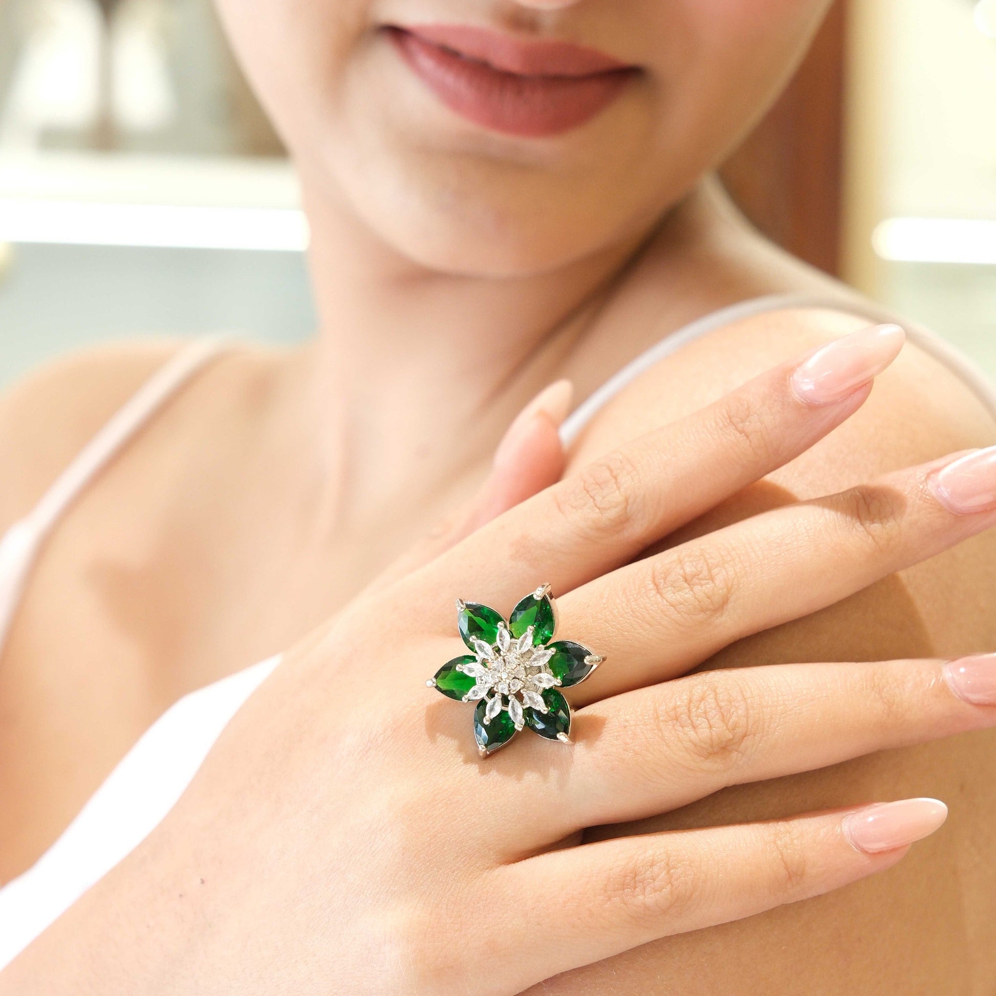 A close-up of a woman's hand wearing a flower-shaped ring with green stones and a silver-toned center, set against her shoulder and upper chest.