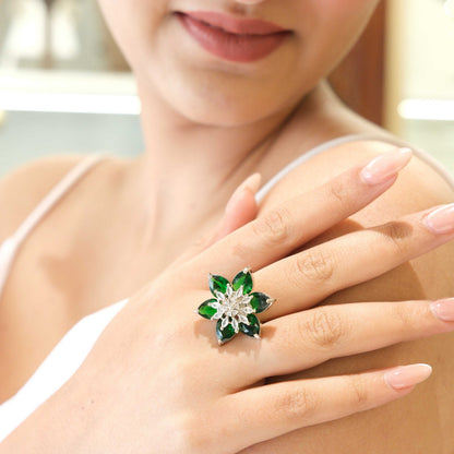 A close-up of a woman's hand wearing a flower-shaped ring with green stones and a silver-toned center, set against her shoulder and upper chest.