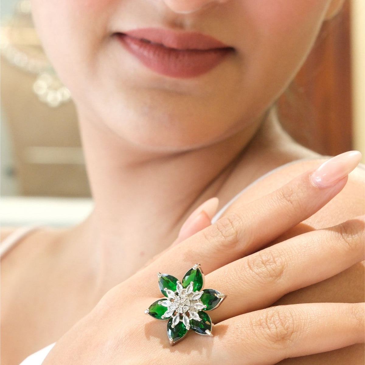 A close-up of a woman's hand wearing a silver-toned flower ring with green gemstone petals and a clear central stone.