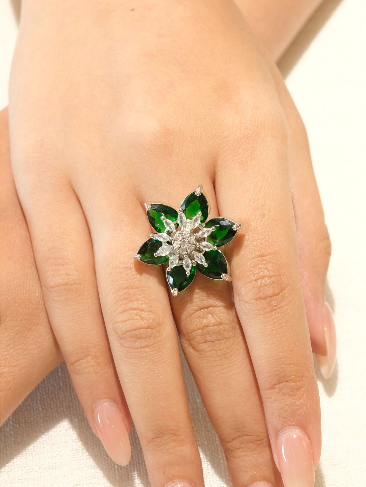 A silver-toned flower ring with green marquise-cut stones and a clear crystal center, shown worn on a hand.