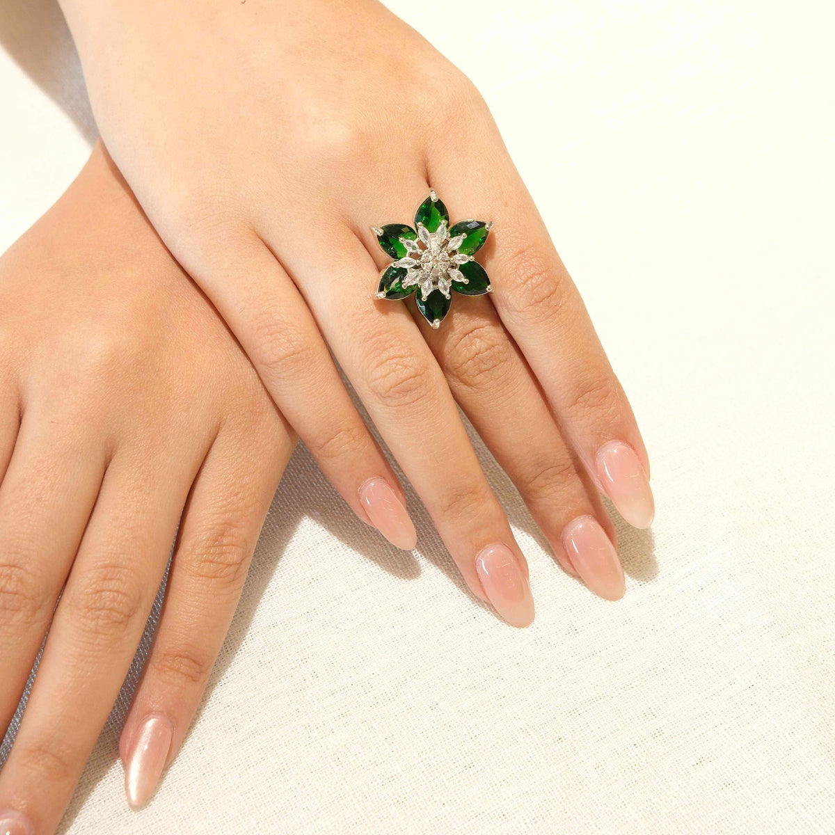 A close-up of a person's hands resting on a white surface, wearing a silver-toned ring shaped like a six-petal flower with green faceted stones and a clear central stone.