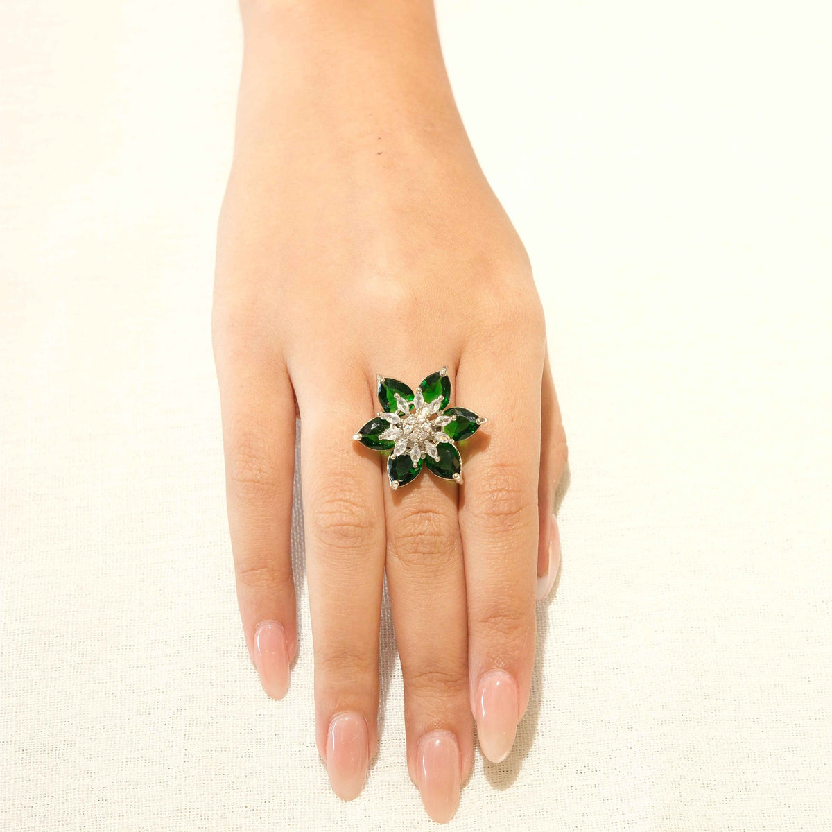 A hand wearing a flower-shaped ring with green stones and a clear central stone, set against a plain off-white background.
