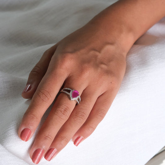 A close-up of a hand wearing a silver ring with a pink heart-shaped gemstone, resting on a white textured fabric background.