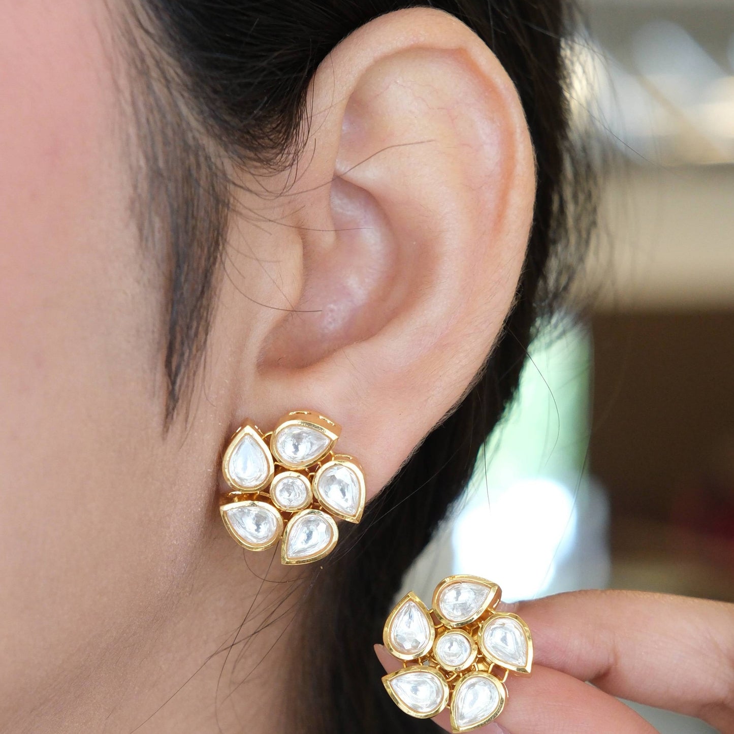 A close-up of a person wearing a gold and white stud earring, with a hand holding the matching earring to show the front design. The earring features a cluster of white teardrop-shaped stones set in a gold-tone circular frame.