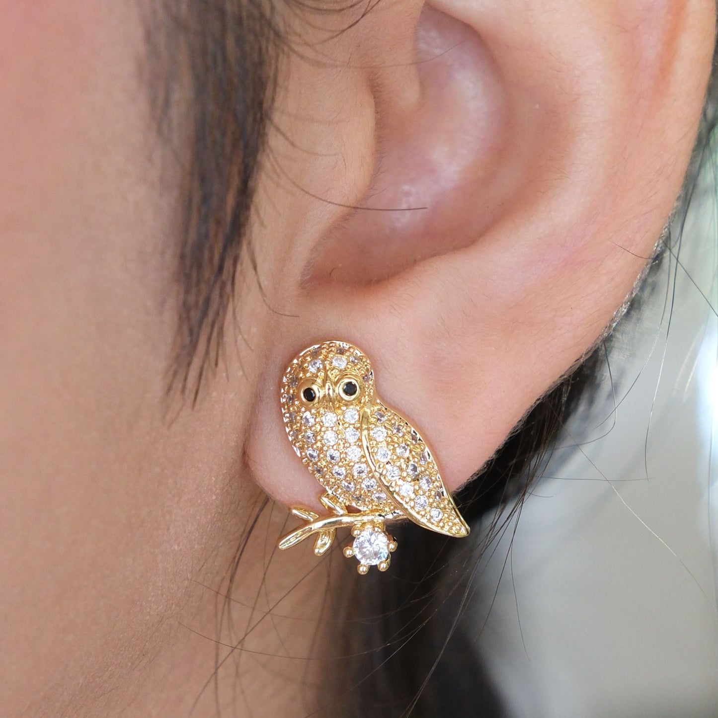A gold owl stud earring paved with small clear crystals, worn on a person's earlobe.
