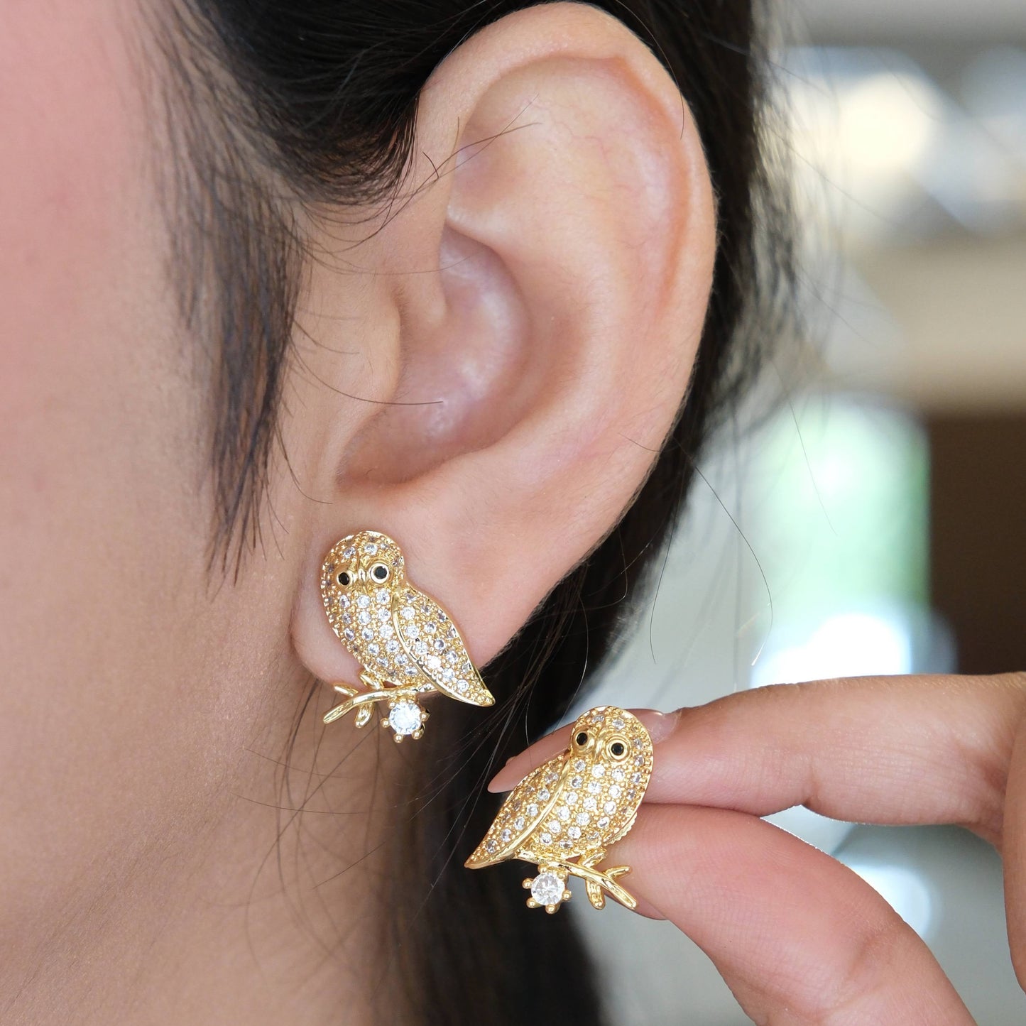 Gold owl stud earrings covered in small clear crystals, shown worn on one ear and held by fingers on the other side.
