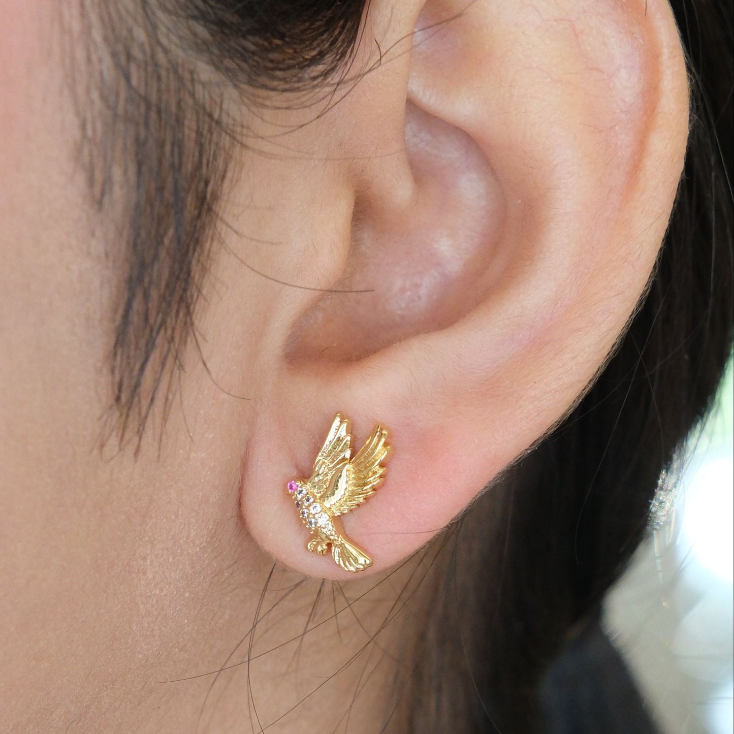 A close-up of a person's ear wearing a small gold bird stud earring with a tiny pink gemstone accent.