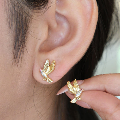 Close-up of a person wearing a small gold bird stud earring on the earlobe, with a hand holding the matching earring against a soft light background.