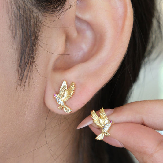 Close-up of a person wearing a small gold bird stud earring on the earlobe, with a hand holding the matching earring against a soft light background.