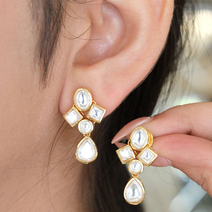 Gold-plated drop earrings with white stones, shown worn on a person's ear and held by a hand.