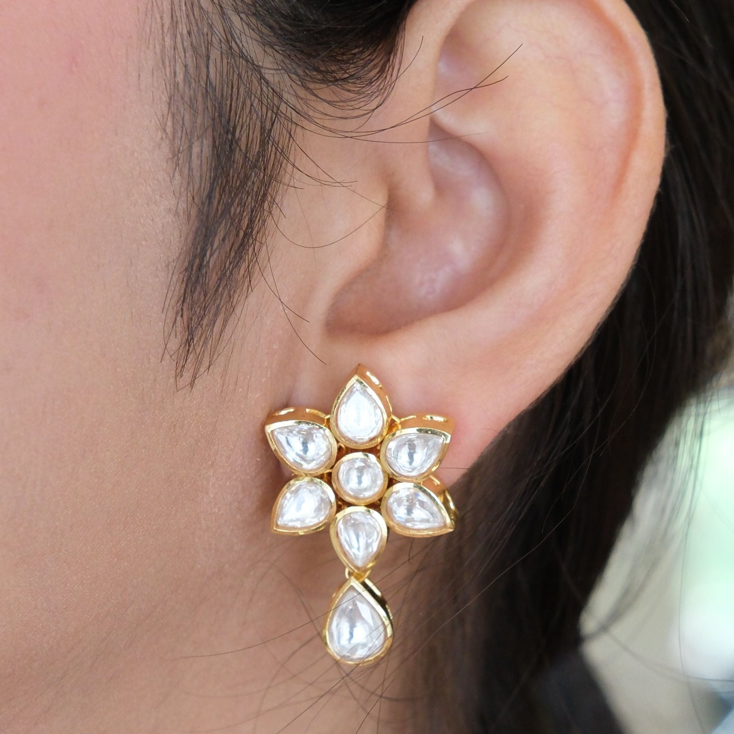 A close-up of a person's ear wearing a gold flower-shaped stud earring with clear teardrop and round stones.