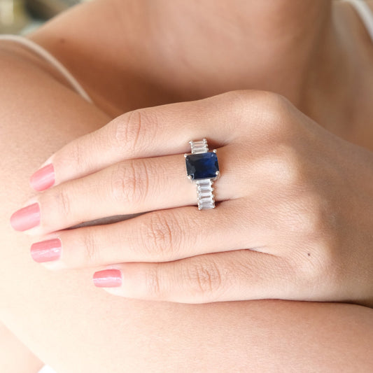 A close-up of a woman's hand wearing a silver ring with a rectangular dark blue gemstone, set against her shoulder and arm.