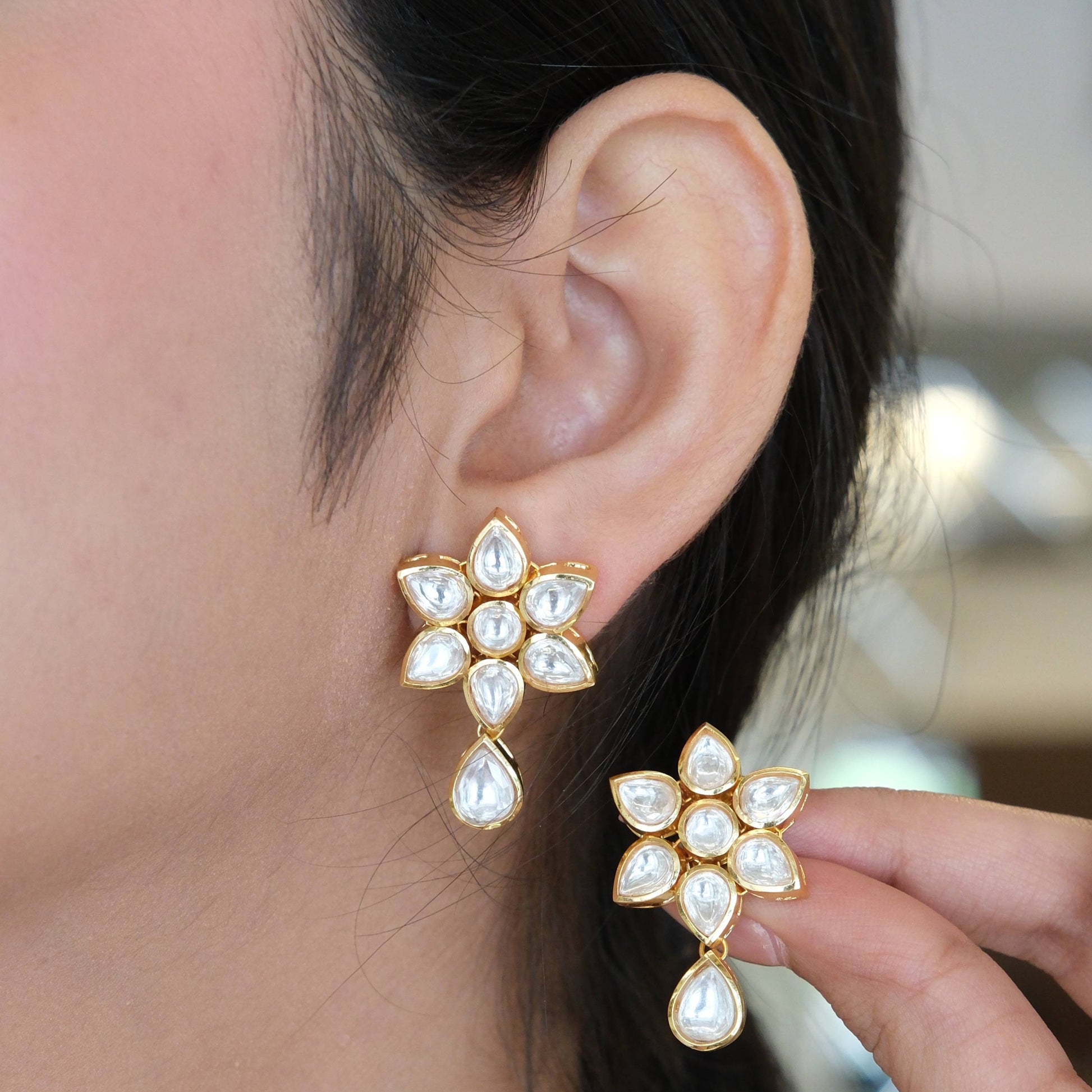 A close-up of a person wearing a gold and white stone flower-shaped dangle earring, with a matching earring held up by a hand.