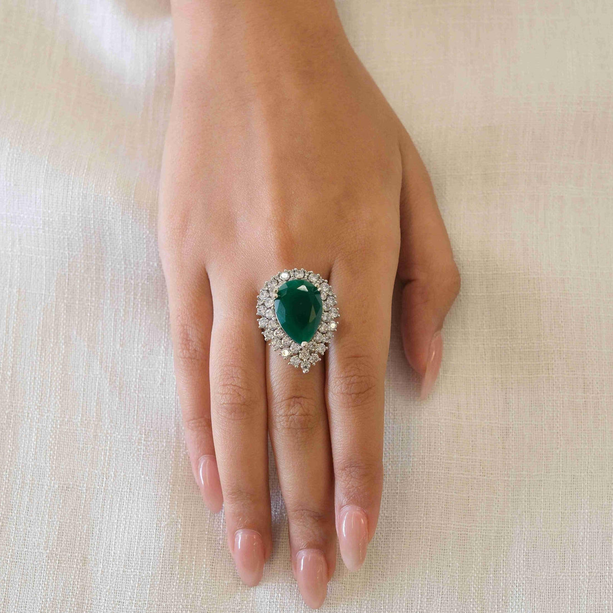 A hand wearing a silver ring with a large oval green emerald center stone and a halo of small clear stones, resting on a light beige fabric background.