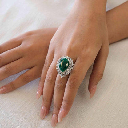 A close-up of a hand wearing a large oval emerald-cut green ring with a halo of small clear stones, resting on a soft cream fabric background.