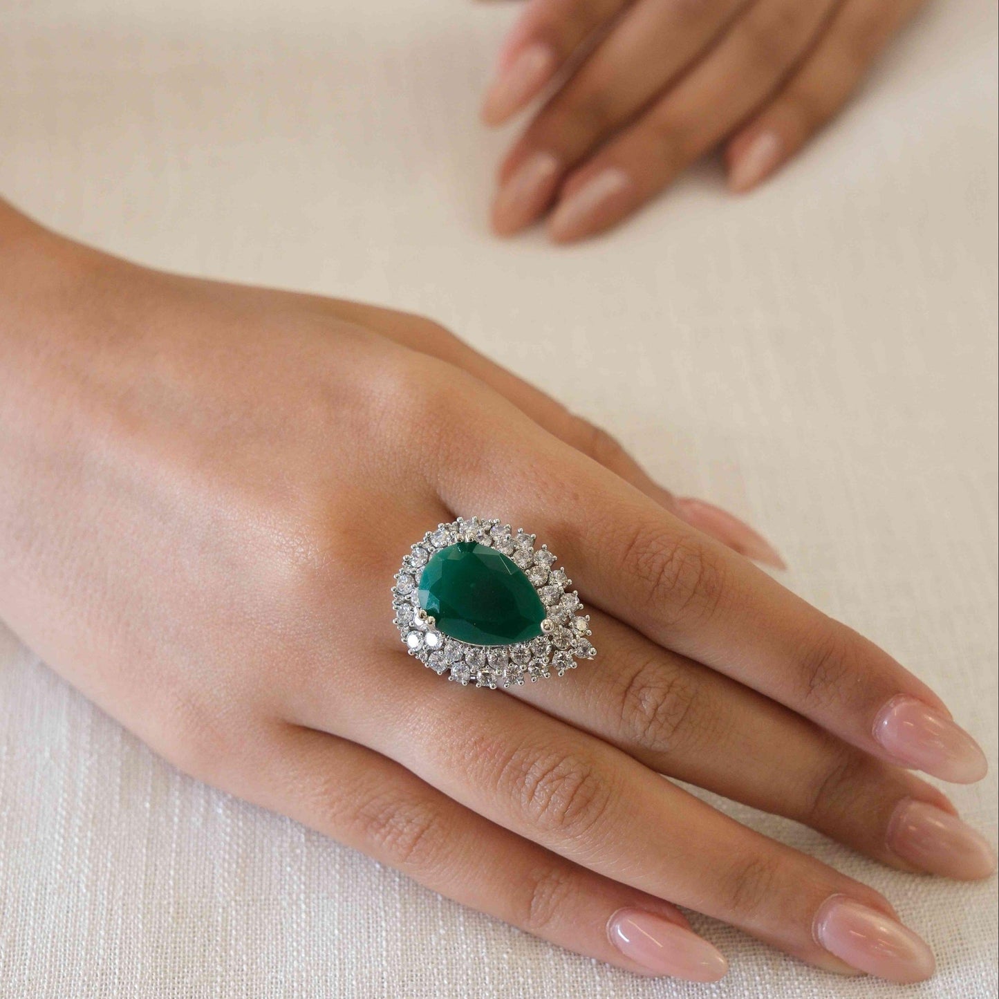 A close-up of a hand wearing a teardrop-shaped ring with a deep green center stone and a halo of small clear stones on a light beige fabric background.