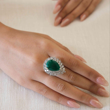 A close-up of a hand wearing a teardrop-shaped ring with a deep green center stone and a halo of small clear stones on a light beige fabric background.