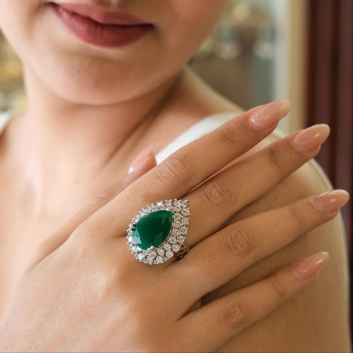 A close-up lifestyle shot of a model wearing a large teardrop-shaped emerald green ring with a diamond halo on her finger.