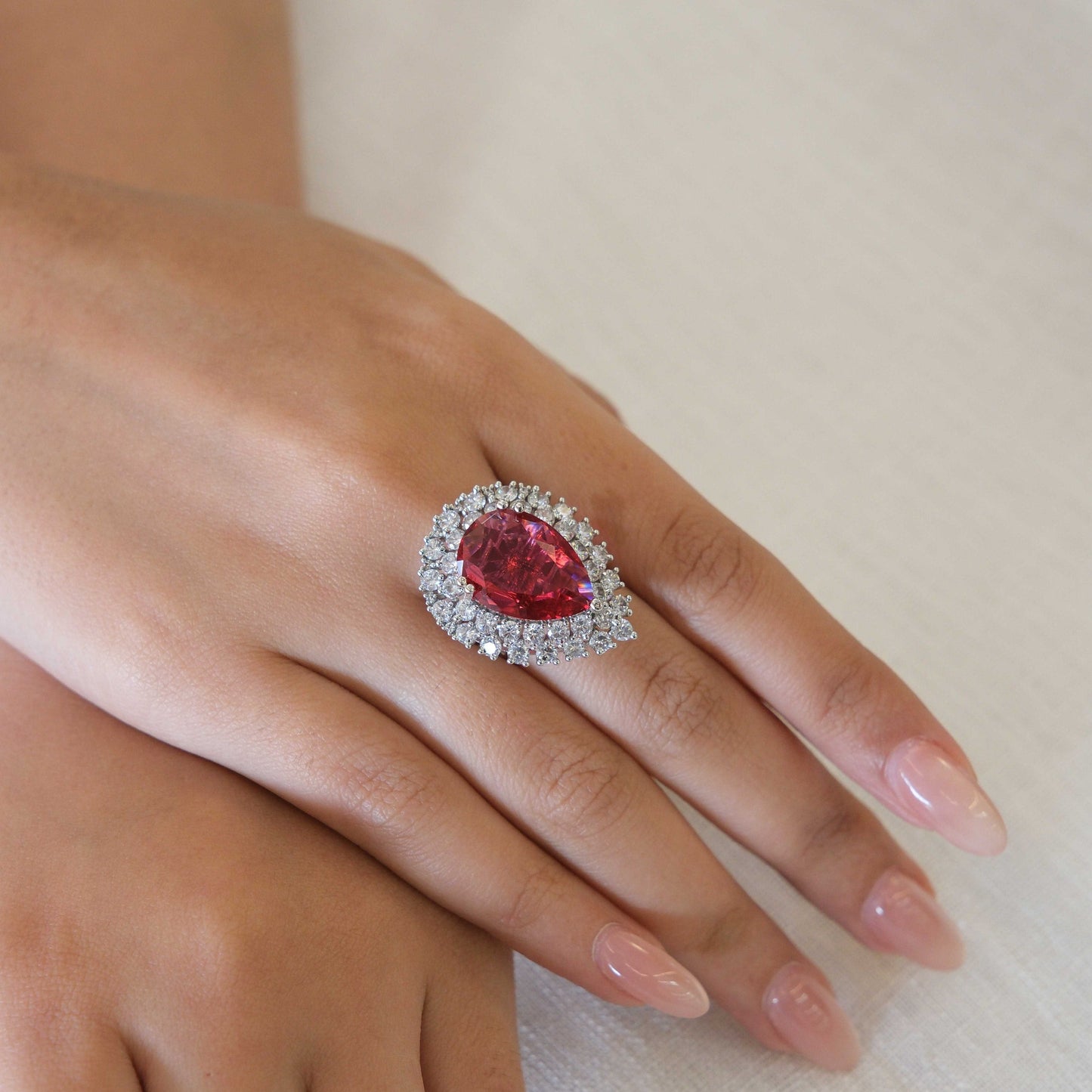 A close-up of a hand wearing a teardrop-shaped red gemstone ring with a silver halo of clear stones, resting on a light beige fabric background.