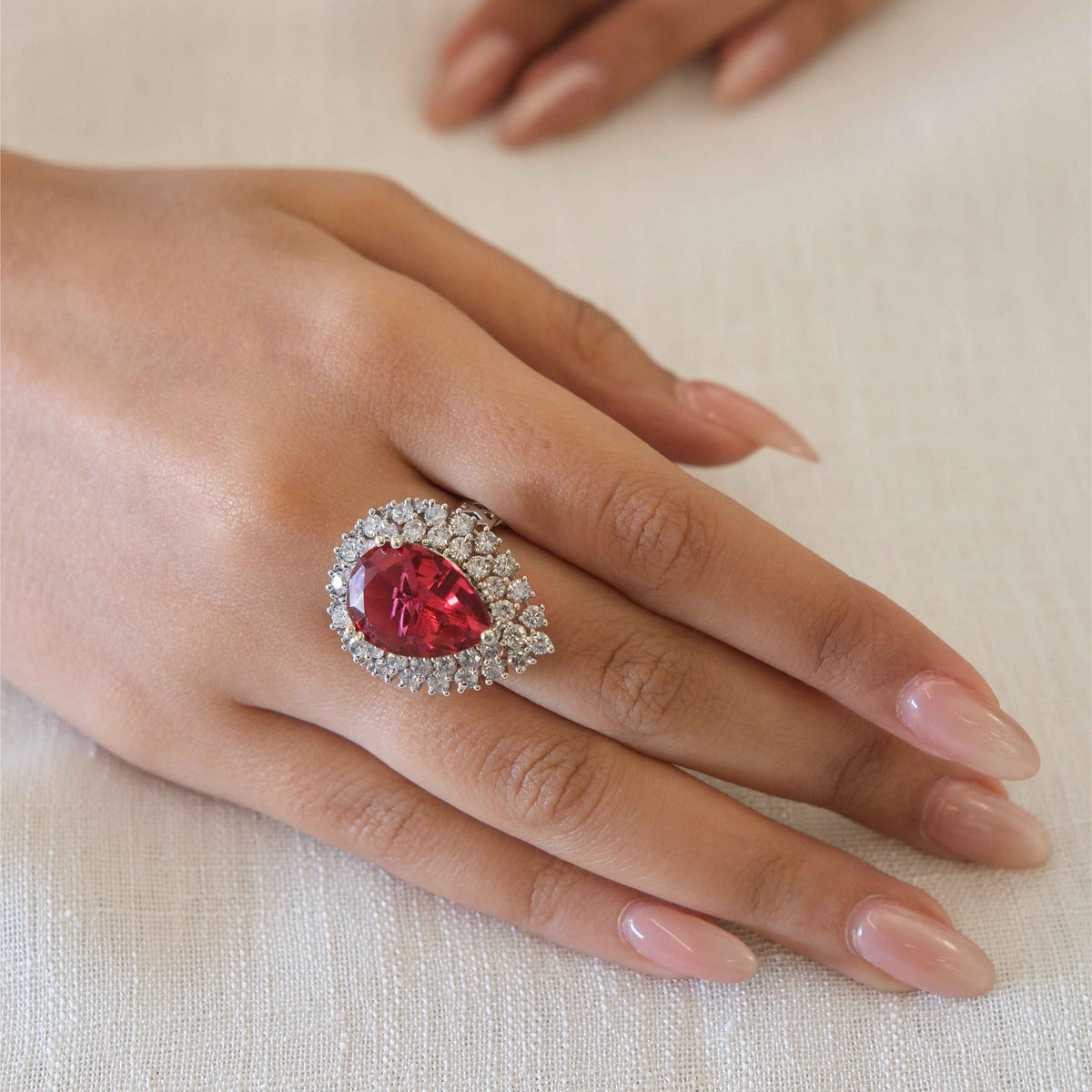 A close-up of a hand wearing a silver ring with a large teardrop-shaped red gemstone surrounded by a halo of small clear stones, resting on a textured off-white fabric surface.