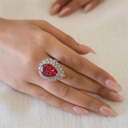 A close-up of a hand wearing a silver ring with a large teardrop-shaped red gemstone surrounded by a halo of small clear stones, resting on a textured off-white fabric surface.