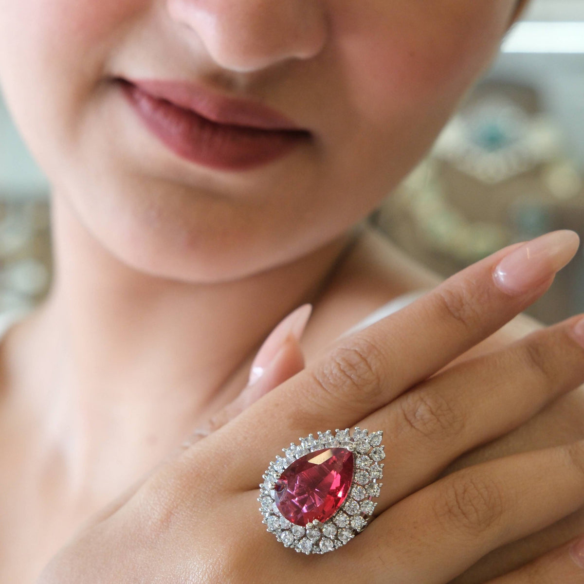 A close-up of a model's hand wearing a large teardrop-shaped pink gemstone ring with a halo of small clear stones.