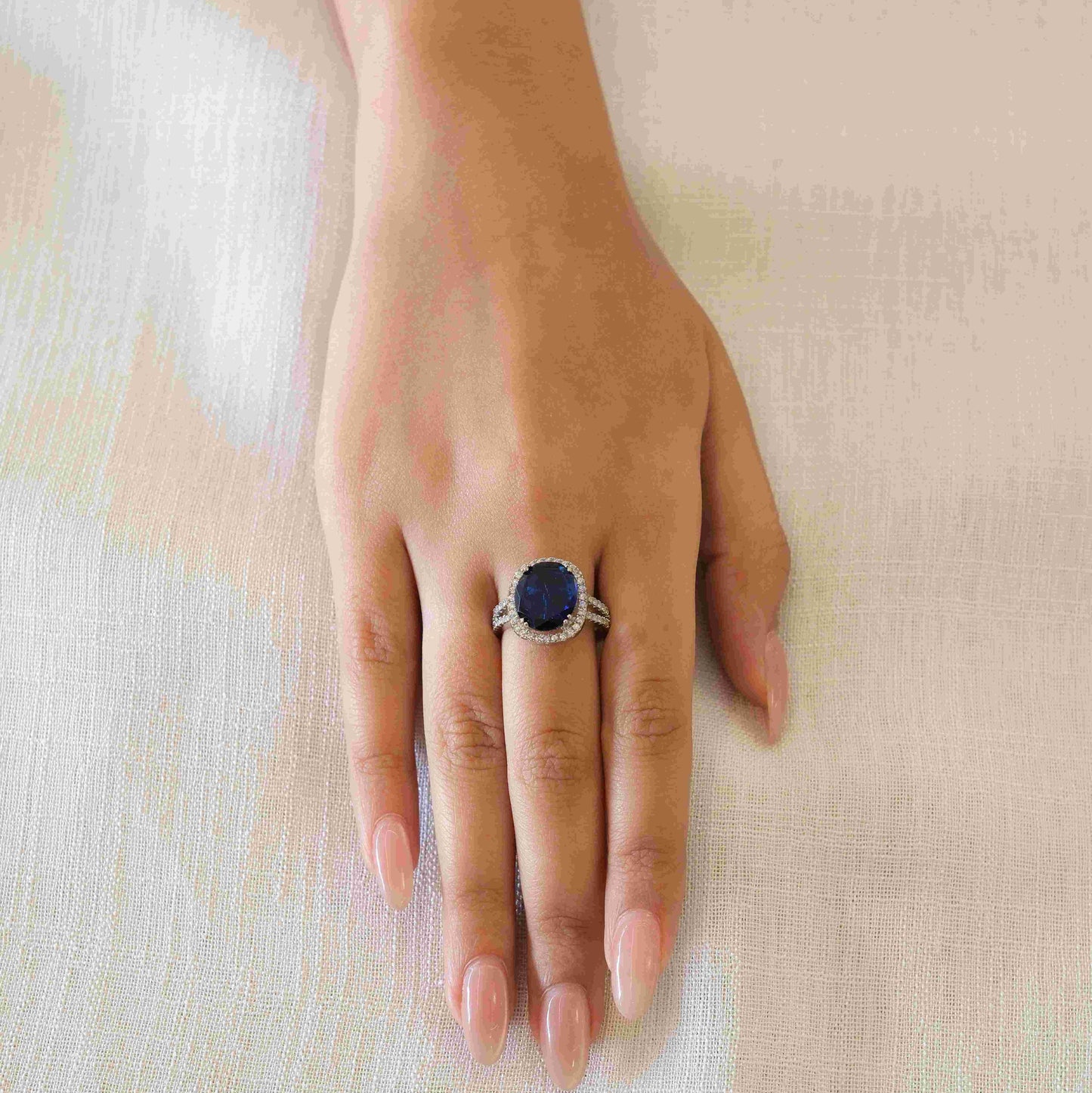 A hand wearing a silver-toned ring with a round dark blue gemstone, resting on a textured beige fabric background.