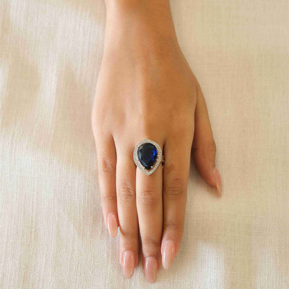 A close-up of a hand wearing a silver ring with a large oval dark blue gemstone, resting on a textured beige fabric background.