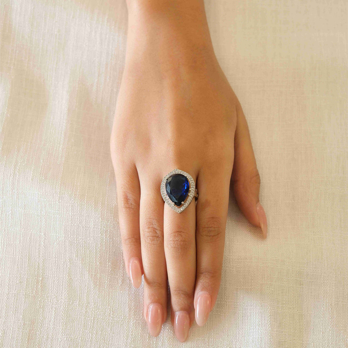 A close-up of a hand wearing a silver ring with a large oval dark blue gemstone, resting on a textured beige fabric background.