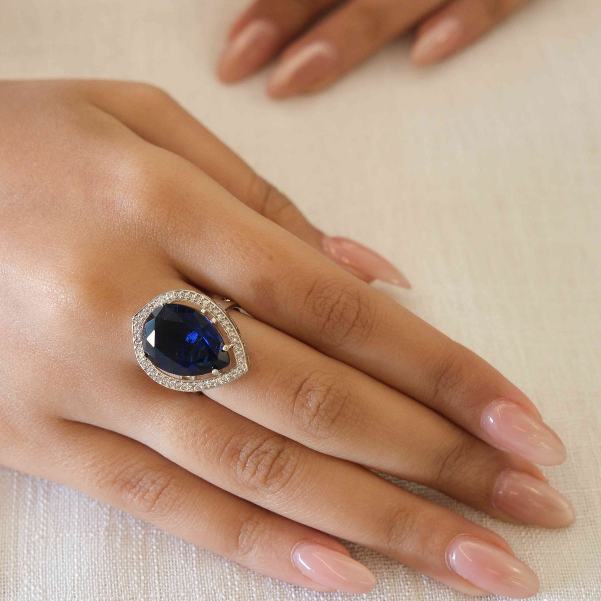 A close-up of a hand wearing a ring with a large oval-cut dark blue gemstone surrounded by a halo of small clear stones, resting on a textured off-white fabric surface.