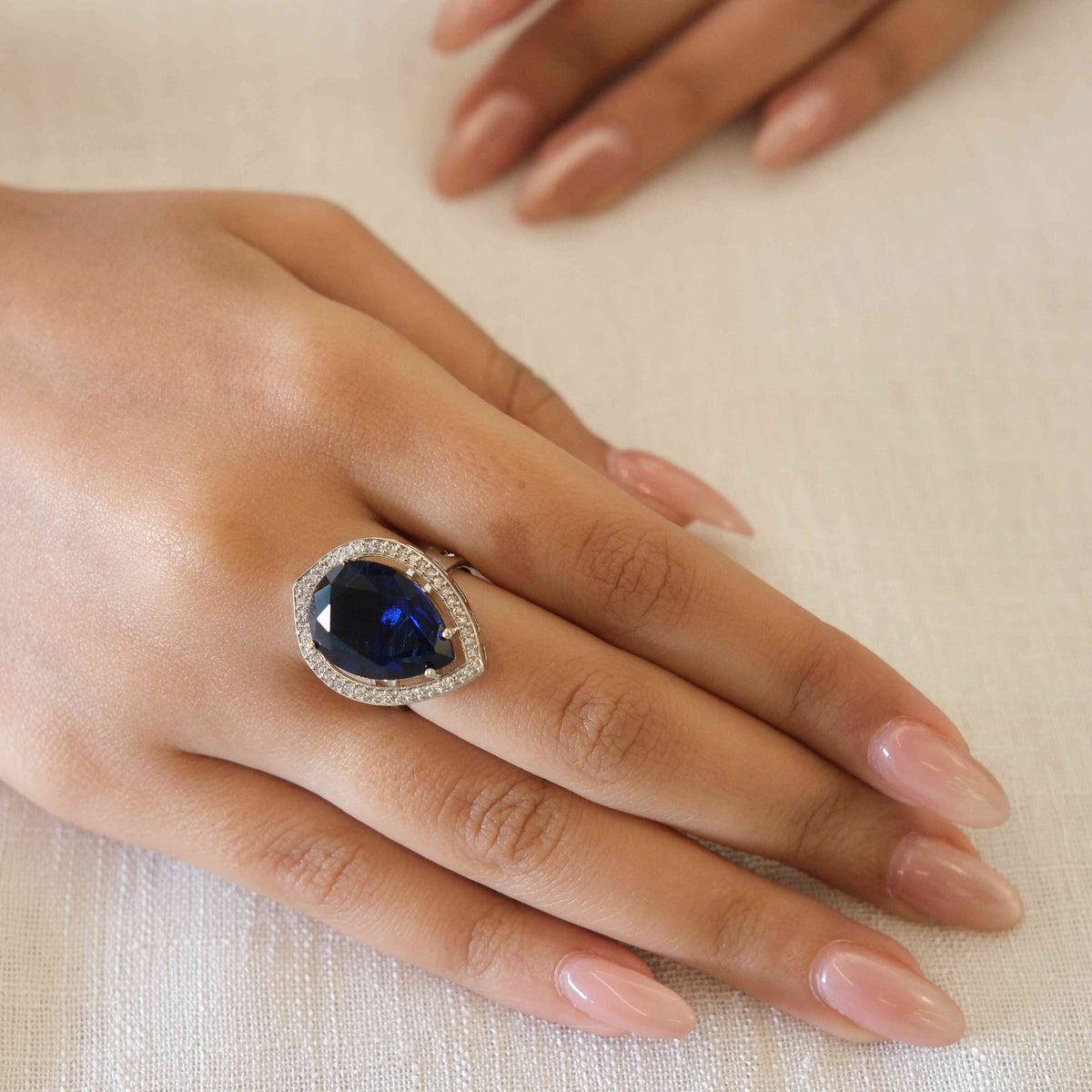 A close-up of a hand wearing a ring with a large oval-cut dark blue gemstone surrounded by a halo of small clear stones, resting on a textured off-white fabric surface.