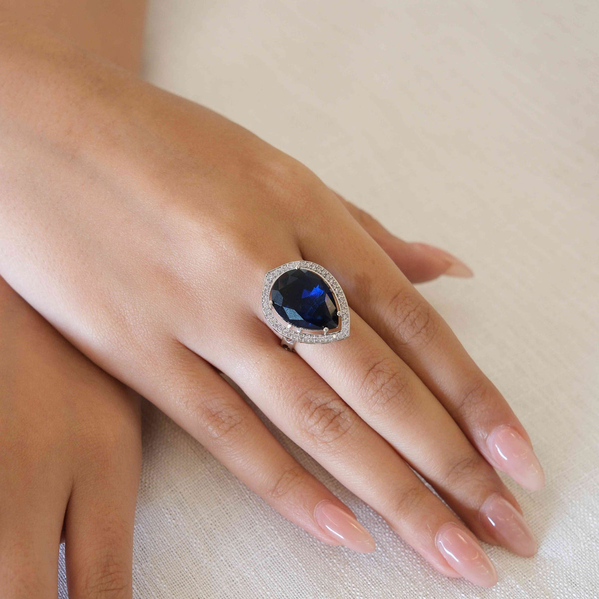 A close-up of a hand wearing a silver-toned ring with a large oval-cut deep blue gemstone surrounded by a halo of small clear stones, resting on a textured off-white fabric background.