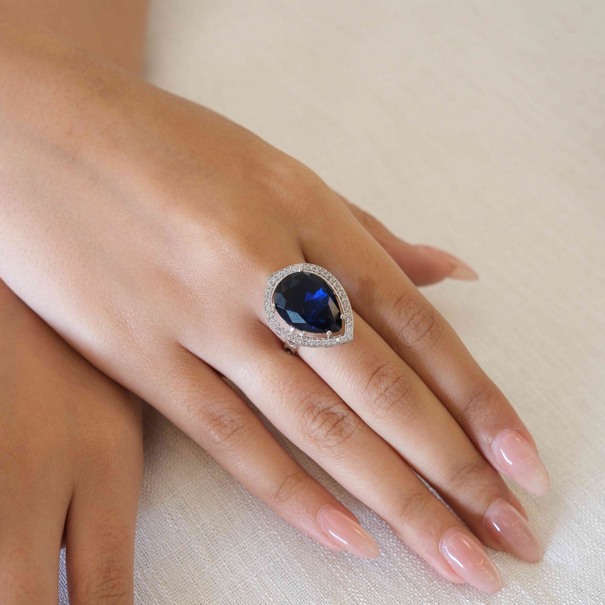 A close-up of a hand wearing a silver-toned ring with a large oval-cut deep blue gemstone surrounded by a halo of small clear stones, resting on a textured off-white fabric background.