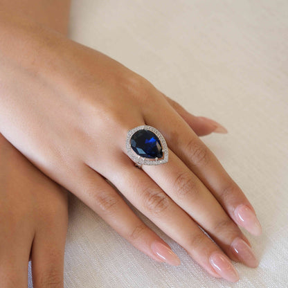 A close-up of a hand wearing a silver-toned ring with a large oval-cut deep blue gemstone surrounded by a halo of small clear stones, resting on a textured off-white fabric background.
