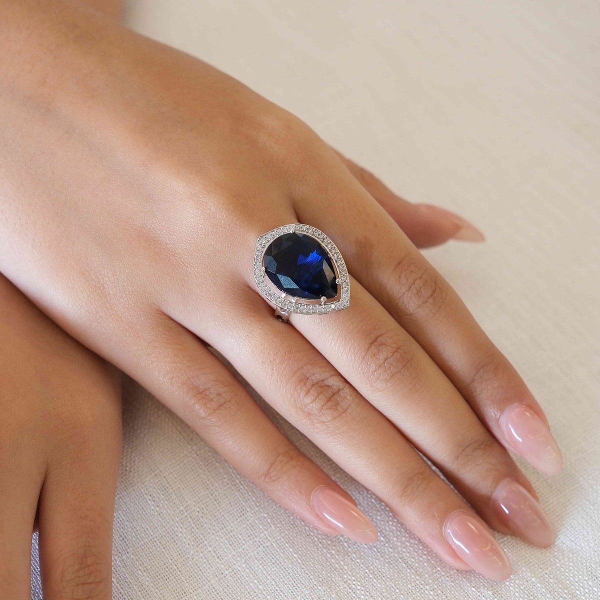 A close-up of a hand wearing a silver-toned ring with a large oval-cut deep blue gemstone surrounded by a halo of small clear stones, resting on a textured off-white fabric background.