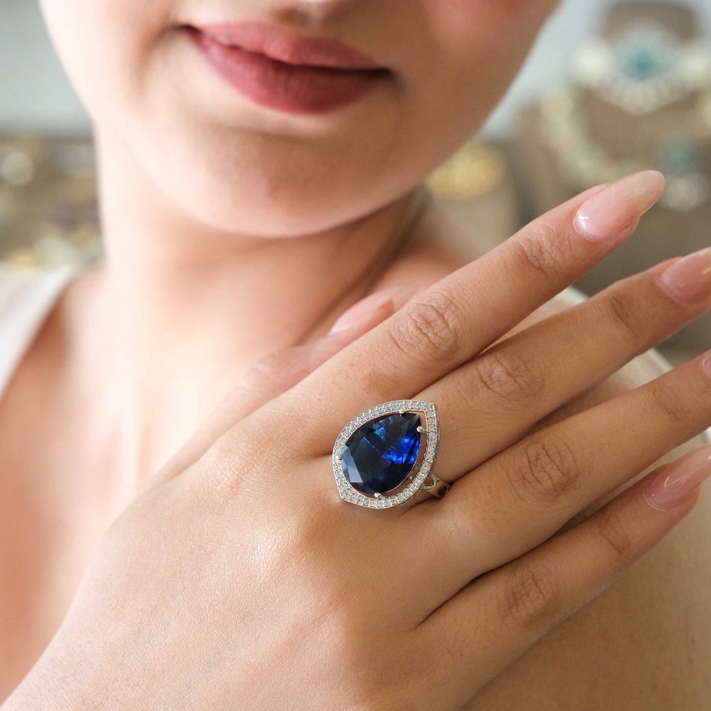 A close-up lifestyle shot of a woman wearing a large teardrop-shaped blue gemstone ring with a halo of small clear stones on her finger.