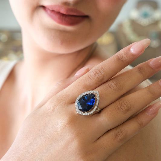 A close-up lifestyle shot of a woman wearing a large teardrop-shaped blue gemstone ring with a halo of small clear stones on her finger.