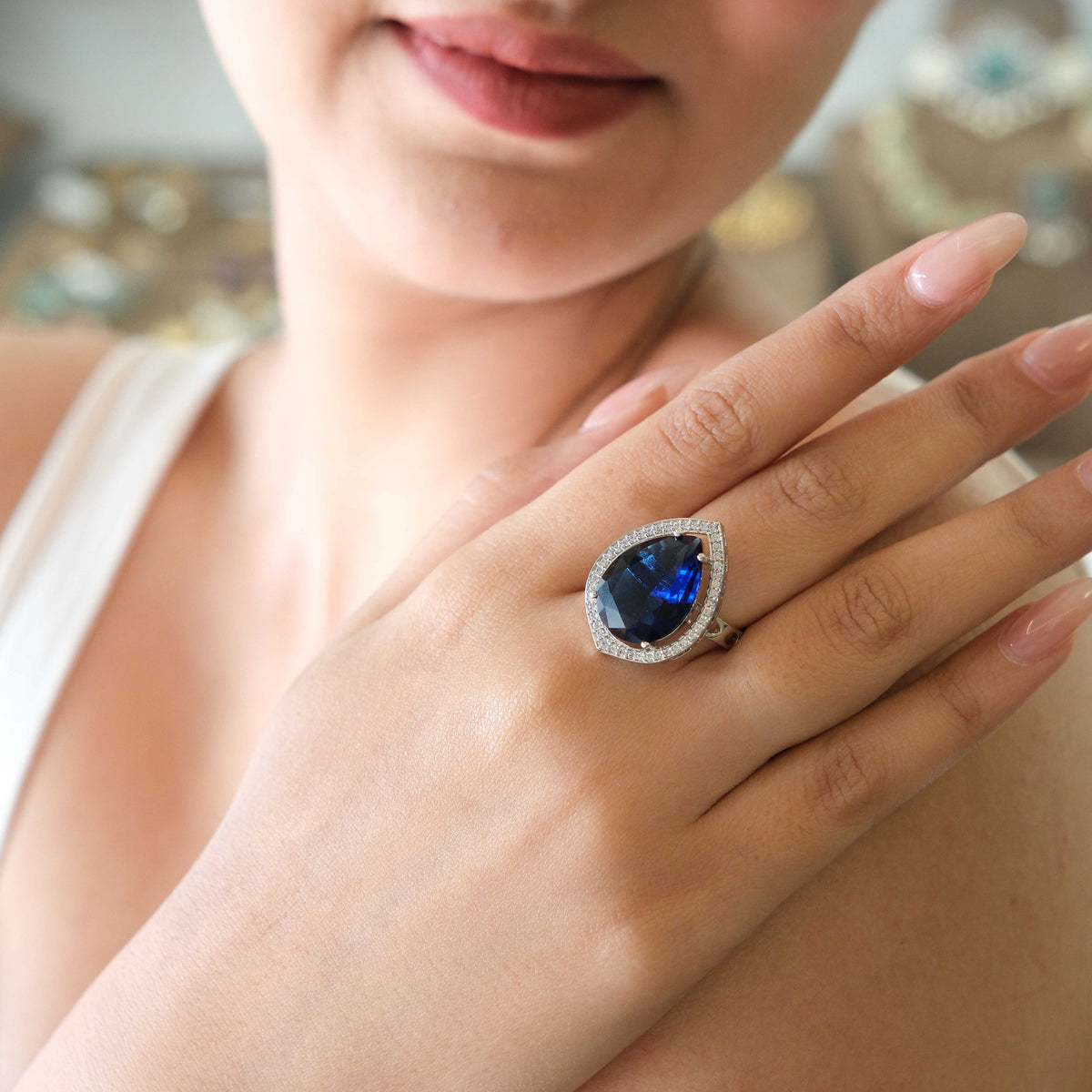 A close-up lifestyle shot of a woman wearing a large teardrop-shaped blue gemstone ring with a halo of small clear stones on her finger.