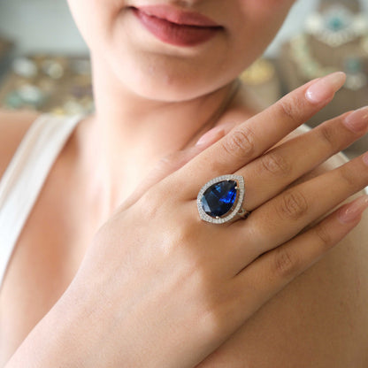 A close-up lifestyle shot of a woman wearing a large teardrop-shaped blue gemstone ring with a halo of small clear stones on her finger.