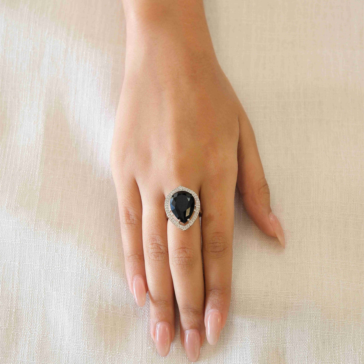 A silver ring with a large black oval stone and a halo of small clear stones, worn on a hand resting on a light beige textured fabric background.